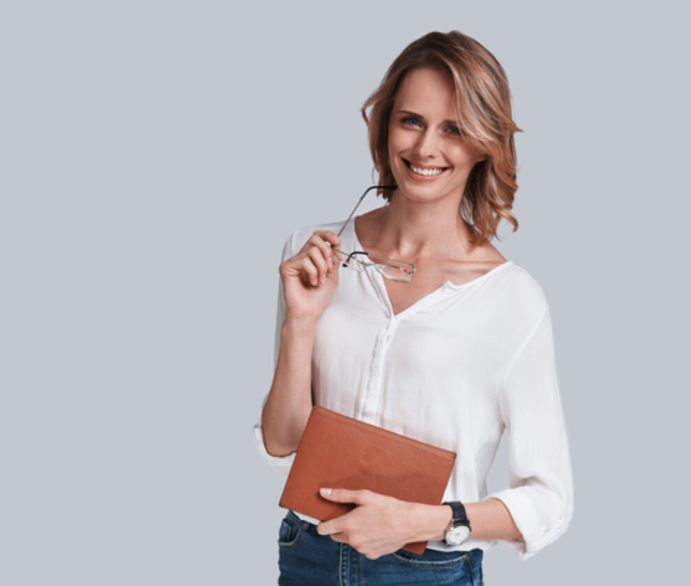 She knows everything about success. Beautiful young woman holding an eyewear and smiling while standing against grey background